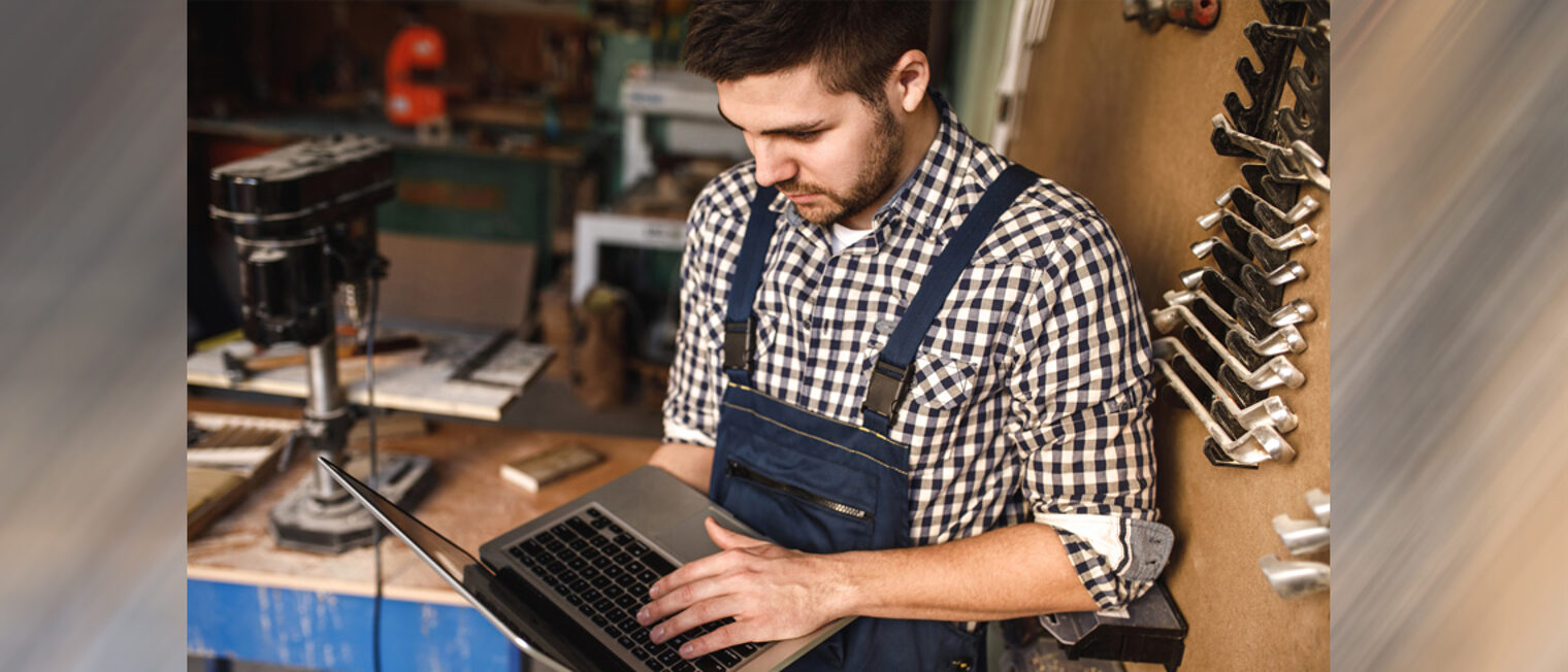 Junger Mann hält einen Laptop in der Hand, er steht in einer Werkstatt