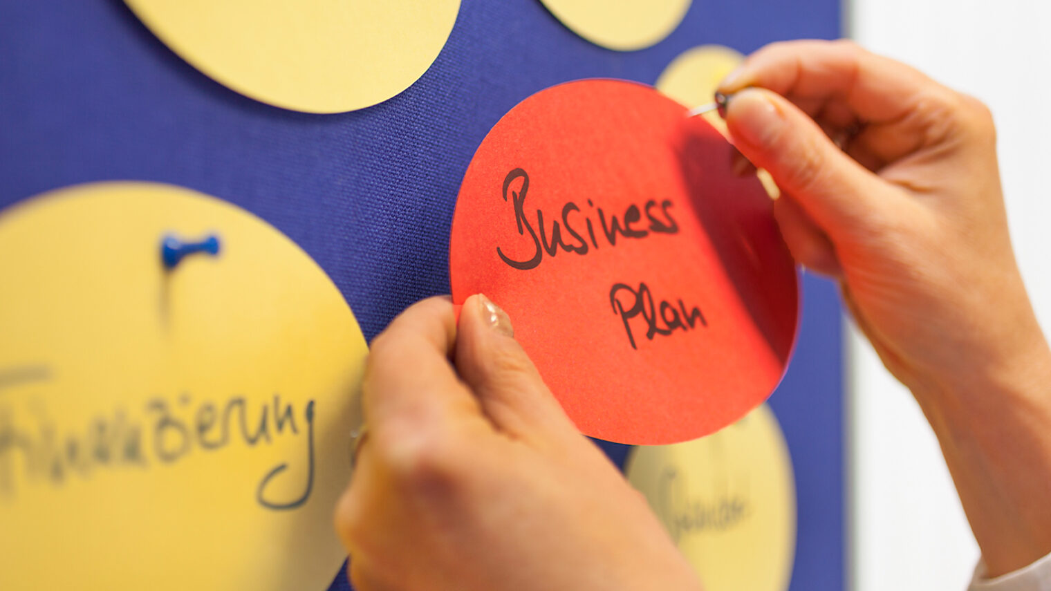 Nahaufnahme von einer Hand, die einen Zettel mit dem Wort Business Plan an eine Wand heftet