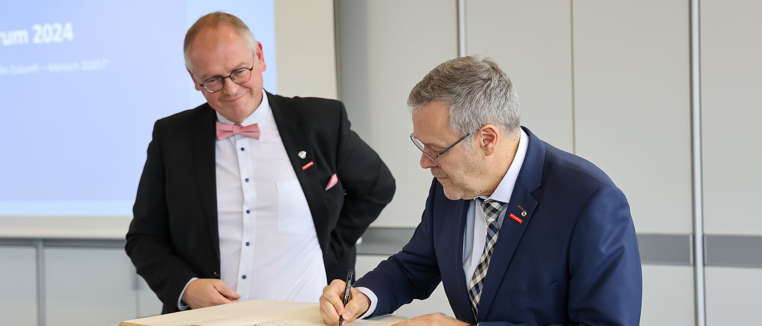 Jörg Dittrich schreibt eine Widmung ins goldenen Buch der Handwerkskammer für Unterfranken. Neben ihm steht Ludwig Paul.