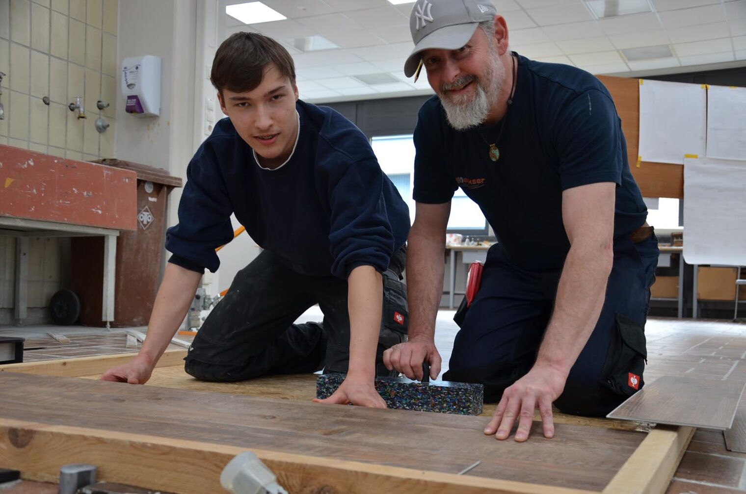 Peter Meeks und Auszubildender Dominik Bund Peter Meeks und Auszubildender Dominik Bund knien auf dem Boden und zeigen das Verlegen von Klick-Vinyl