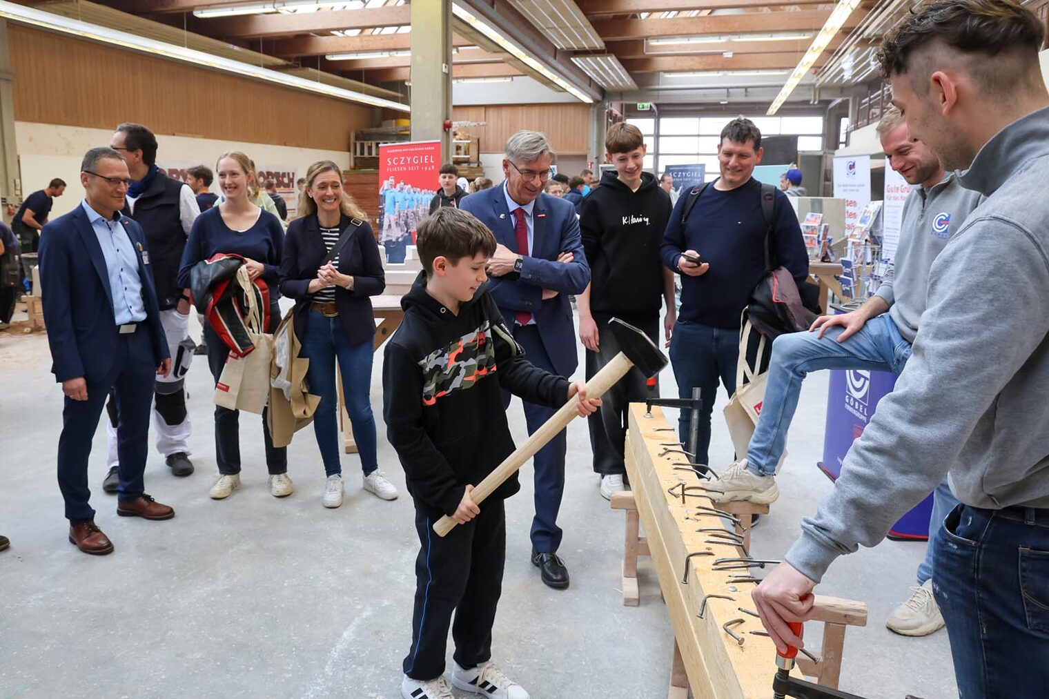Hammer-Aktion beim Macher-Tag 2025 Besucher des Macher-Tags schlägt langen Nagel mit großem Hammer in Holz-Balken vor Publikum, das im Kreis steht.