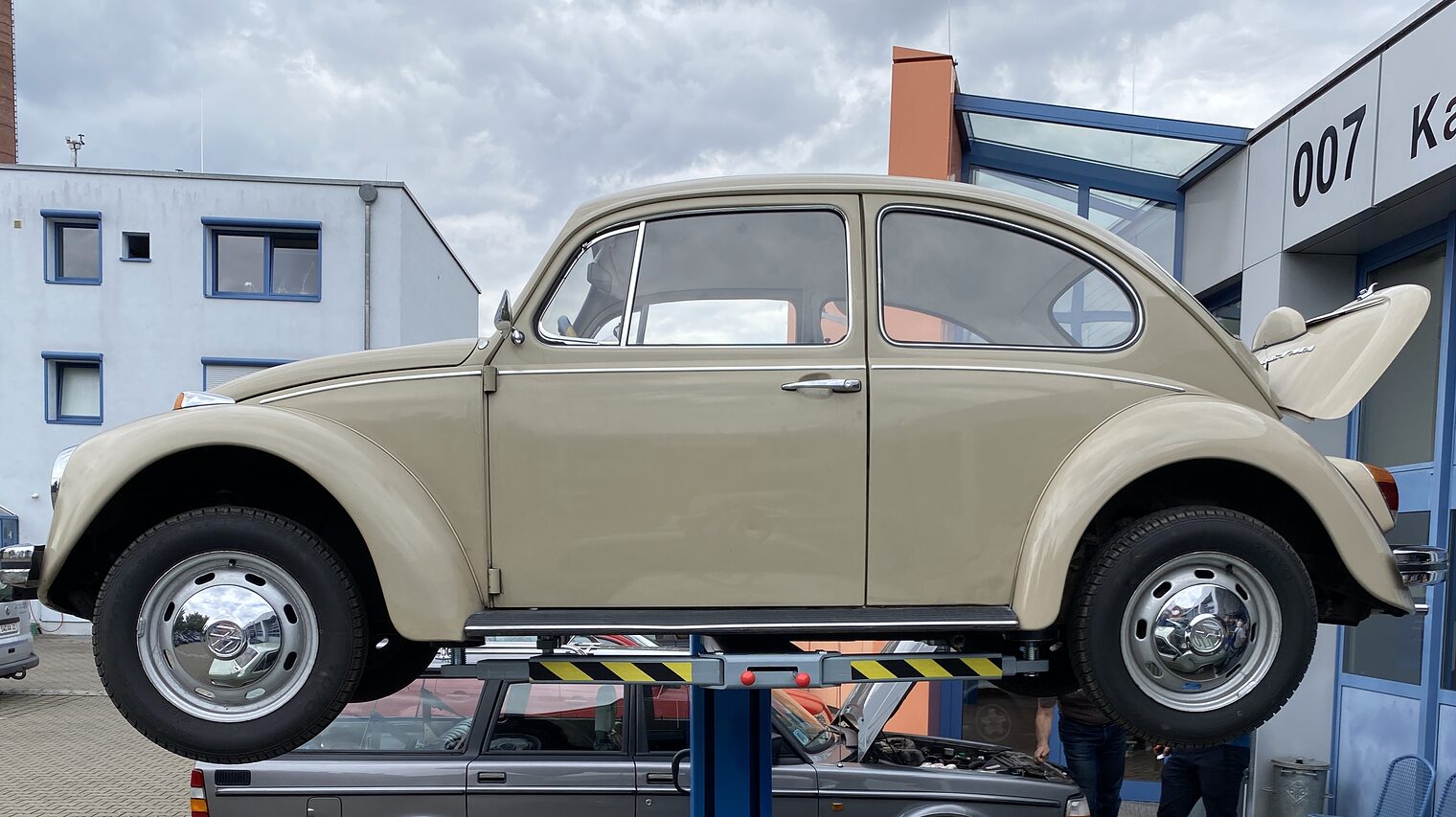 VW Käfer Beiger VW Käfer steht auf Hebebühne vor Werkstatt, darunter grauer Volvo, zwei Personen arbeiten am geöffneten Motorraum.