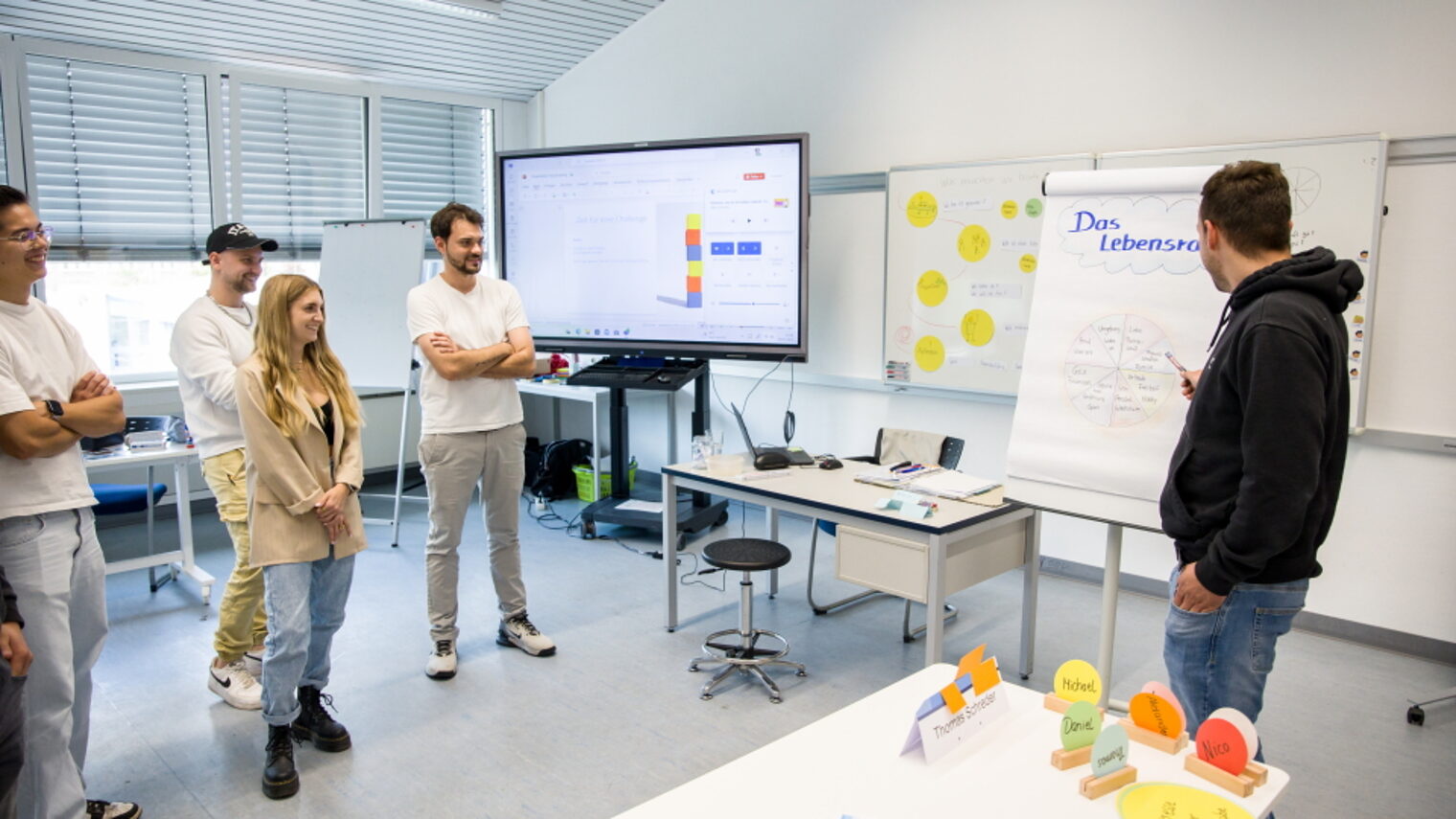 Geprüfter Betriebswirt HwO Schwerpunkt Fahrzeugwesen Eine Gruppe junger Menschen steht im Seminarraum, ein Mitschüler erklärt am Flipchart "Das Lebensrad“.