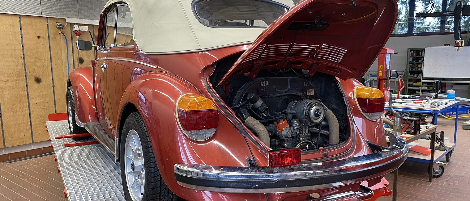 Ein roter VW Käfer mit offenem Motorraum steht in einer Werkstatt auf einer Hebebühne.
