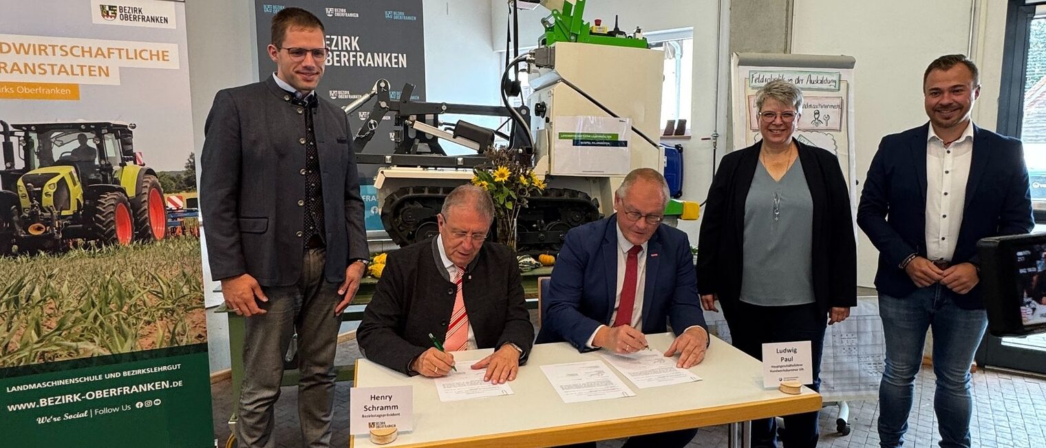 Unterschrieben zum Start des Projektes gemeinsam den Kooperationsvertrag Fünf Personen sind auf dem Bild. Zwei sitzen am Tisch und unterschreiben einen Vertrag. Drei Personen stehendrumherum