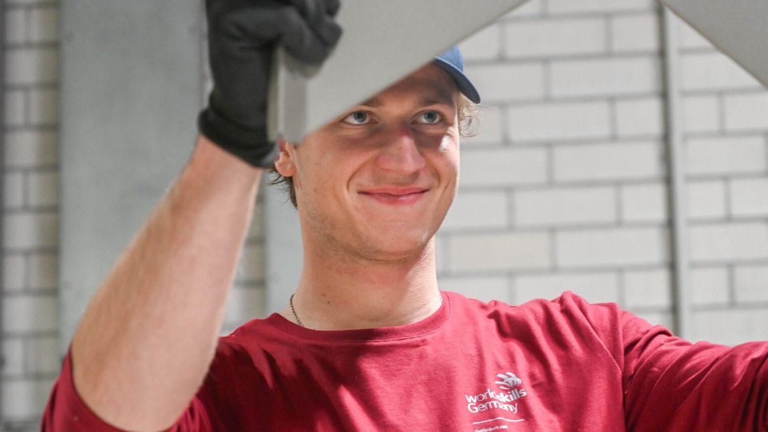 Junger Mann in rotem Oberteil (WorldSkills Logo) hält mit Arbeitshandschuhen ein Blech in die Höhe