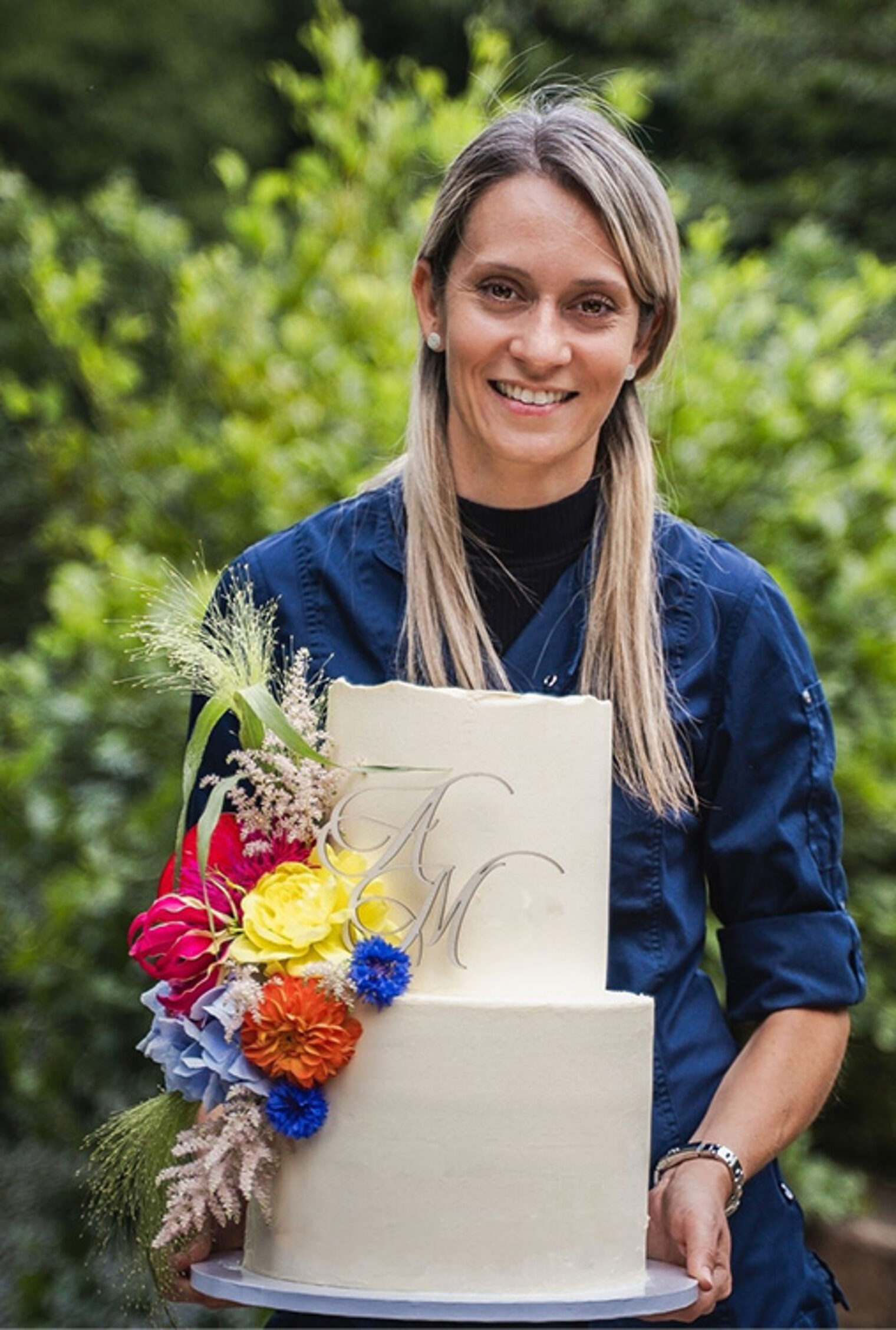 Die Dozentin Mona Mocciaro steht in der Natur vor einem Busch und hält lächelnd eine Torte in ihren Händen.