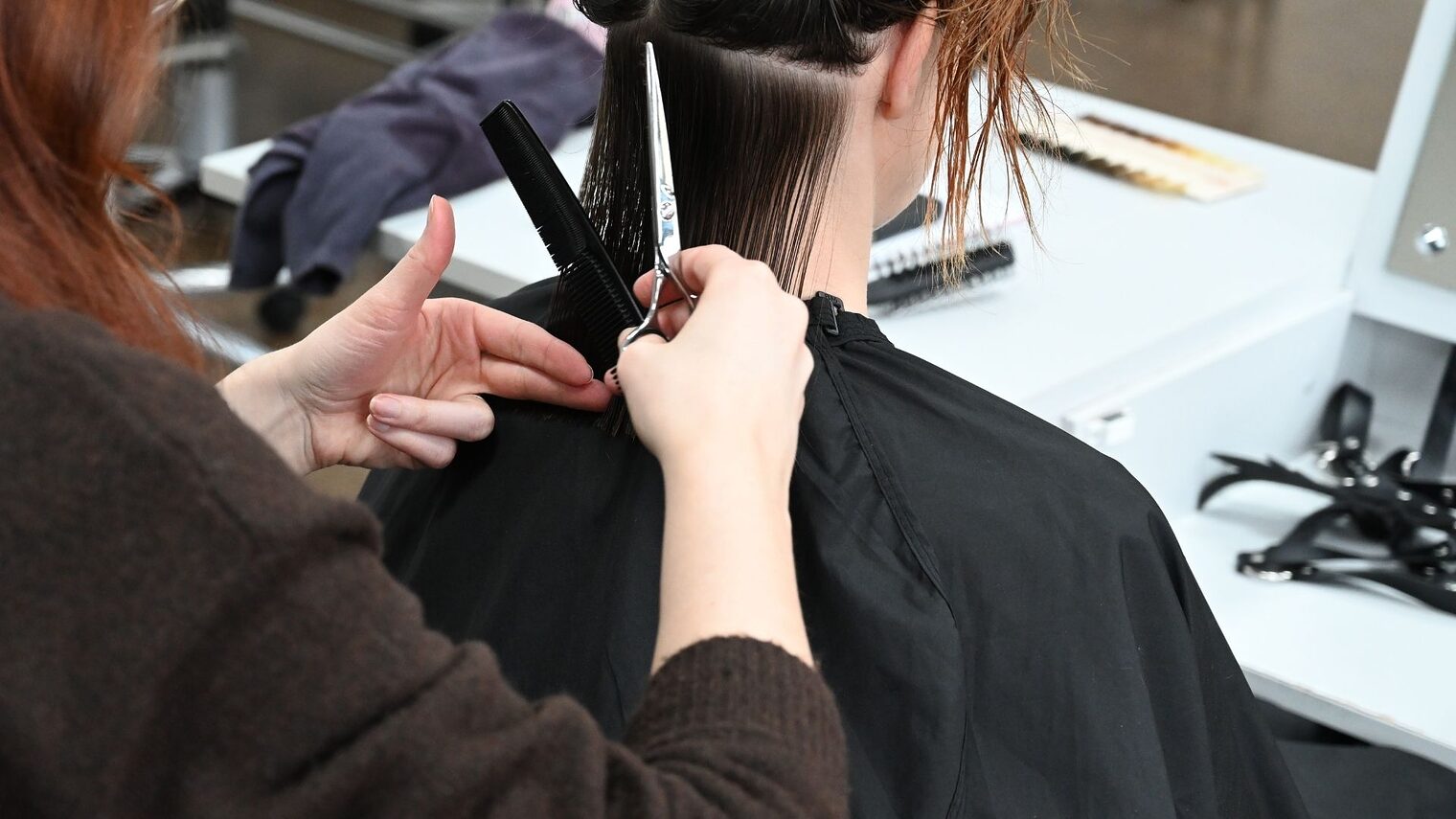 Situation von hinten fotografiert - eine Friseurin mit Kamm und Schere in der Hand schneidet einer sitzenden Dame die Haare