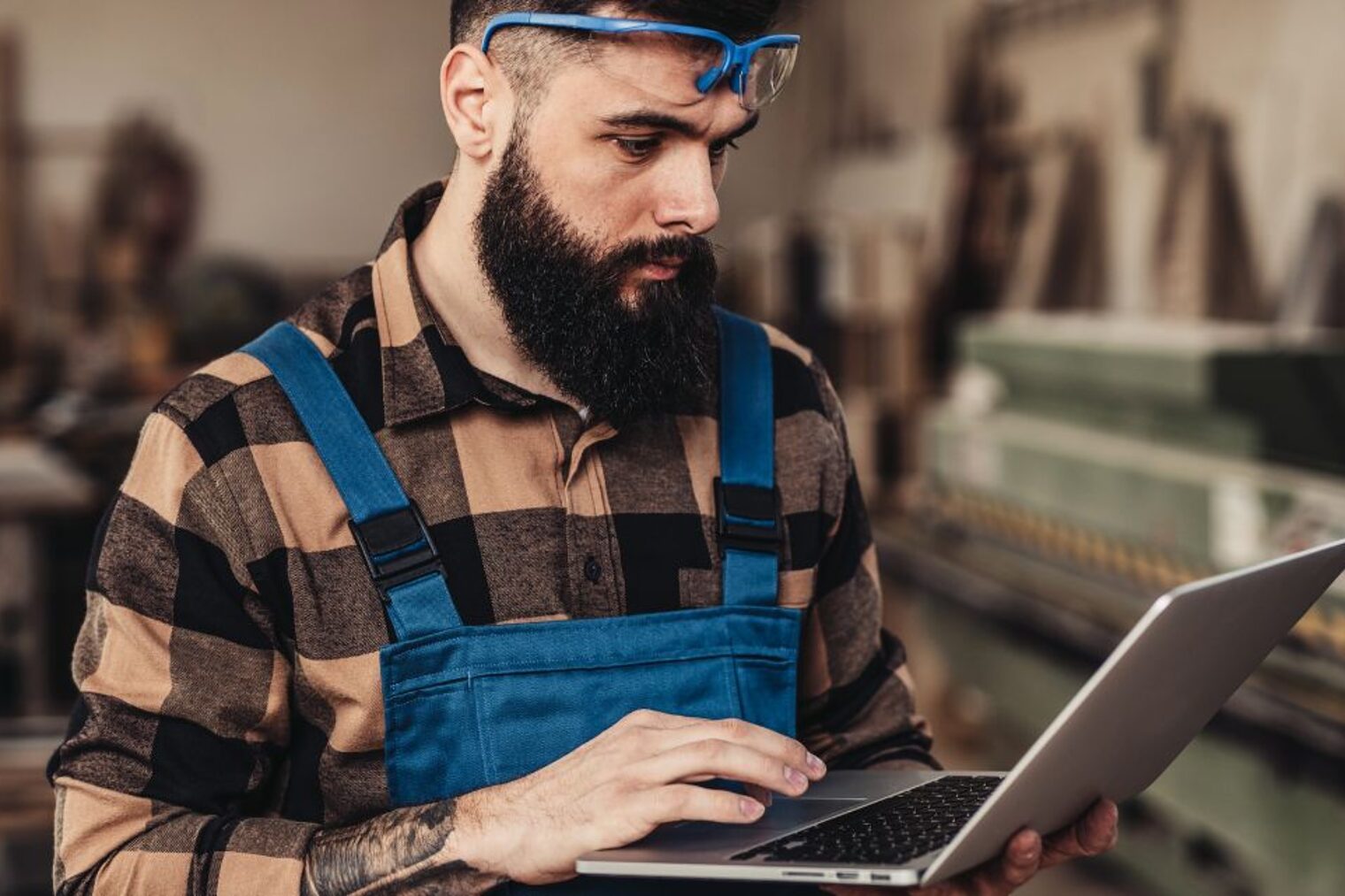 Mann mit kariertem Hemd und blauer Latzhose steht in einer Werkstatt und schaut auf einen Laptop, den er in den Händen hält