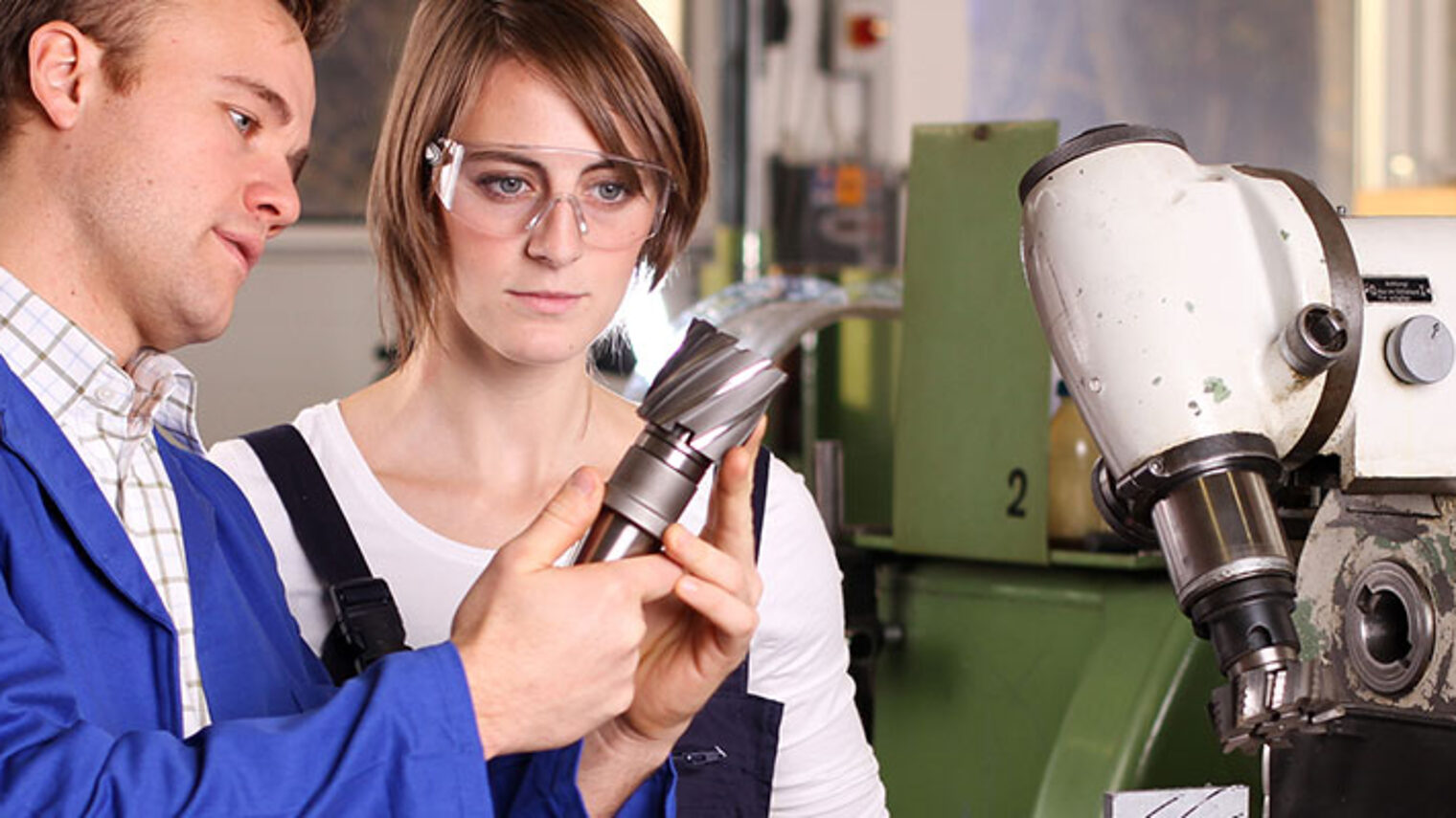 Mann zeigt Auszubildender ein gedrehtes Metallteil in der Werkstatt vor einer Maschine